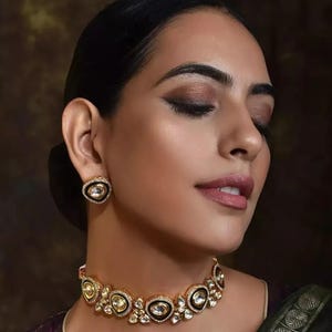 May include: A gold necklace and matching earrings set. The necklace and earrings feature oval, clear gemstones set in a gold frame with black accents. The earrings are round and the necklace sits around the neck.