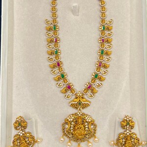 May include: A gold-toned necklace and earring set featuring intricate metalwork, embedded with rectangular green and pink stones, small white accents, and a central pendant depicting a deity figure, finished with dangling white pearl-like beads.