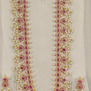 May include: A gold-coloured jewellery set comprising a necklace and earrings. The necklace and earrings are decorated with ruby-coloured rectangular and floral-shaped stones, and small white pearls. The set is displayed against a neutral background.