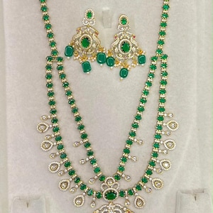 May include: A jewelry set consisting of a long, two-strand necklace and matching earrings. The pieces feature gold-tone metal, oval-cut green stones, and clear white stones. The necklace has a large central pendant with dangling green stones.