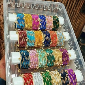 May include: A collection of multi-colored metal bangles arranged on four white cylindrical holders inside a clear plastic storage box. Each bangle features a textured surface with small gold-toned decorative accents attached throughout.