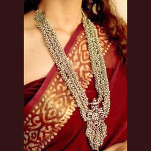 May include: A silver necklace with multiple strands of beads and a large pendant. The pendant is ornate and has a floral design. The necklace is worn by a person with a red and gold patterned saree.