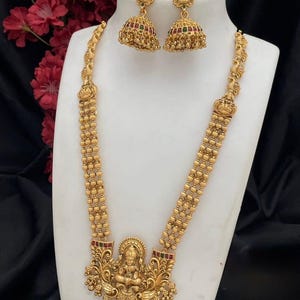 Könnte beinhalten: Goldfarbenes Halsketten- und Ohrring-Set, präsentiert auf einer weißen Büste. Die Halskette hat einen detaillierten Anhänger mit einer zentralen Figur und aufwendigen Verzierungen. Die Ohrringe sind glockenförmig mit roten und grünen Akzenten. Das Set ist aufwendig gestaltet.