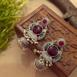 May include: A pair of silver-toned dangle earrings featuring a large circular deep red stone, a smaller red stone at the top, and green accents. The earrings include intricate metalwork and small white beads hanging from a bell-shaped base.