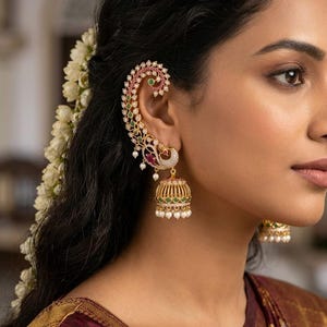 Può includere: Un orecchino a cerchietto dorato con motivo a pavone decorato con piccole pietre bianche, accenti rosa e verdi, e un pendente a forma di campana jhumka impreziosito da pietre verdi e piccole perle bianche pendenti dalla base.