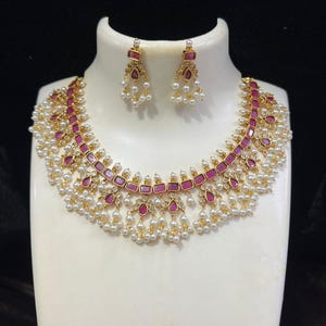 Könnte beinhalten: Eine goldene Halskette mit rosa und weißen Steinen und ein passendes Paar Ohrringe. Die Halskette hat eine Reihe von rosa Steinen mit weißen Steinen dazwischen und einen Fransen aus weißen Perlen.
