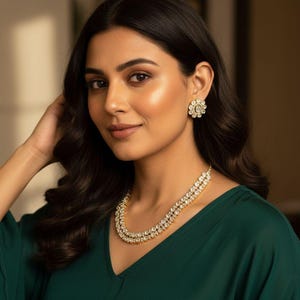 May include: A woman wearing a gold necklace and matching earrings. The necklace features two rows of sparkling stones. The earrings are round with a floral design. She is wearing a dark green top.