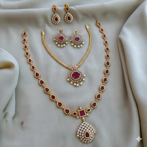 Op de afbeelding: Een goudkleurige sieradenset met een ketting, oorbellen en een armband. De ketting en armband hebben druppelvormige rode edelstenen. De oorbellen en hanger hebben een centrale rode steen omringd door kleinere witte stenen en parels.