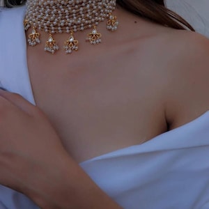 Könnte beinhalten: Eine mehrreihige Perlenkette mit goldenen Akzenten und herabhängenden Perlentropfen. Die Kette besteht aus kleinen weißen Perlen und hat ein zartes, feminines Design.