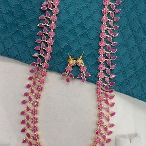 Puede incluir: Un conjunto de joyas dorado con un collar y pendientes a juego. El collar y los pendientes están adornados con piedras preciosas rosas en forma de lágrima y pequeños detalles florales. El conjunto se exhibe sobre un fondo de tela turquesa.