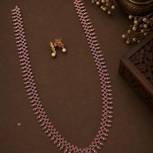 May include: A long gold-finish necklace featuring a repeating pattern of small, teardrop-shaped pink stones and a single white pearl drop at the center, accompanied by a pair of matching stud earrings with pink stones and white pearl accents.