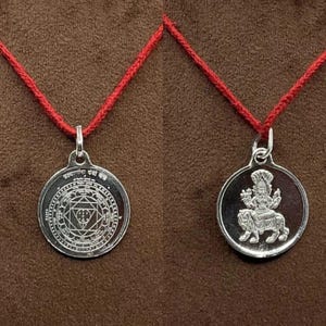 May include: Two silver pendants on red cords. One pendant features geometric patterns and text, the other depicts a deity riding an animal. The pendants are round and have a shiny finish.