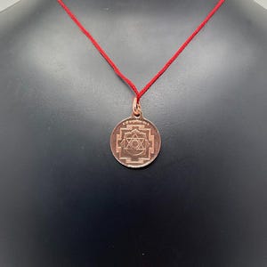 May include: A copper pendant necklace with a red string. The pendant features a circular design with a star and an eye in the center.