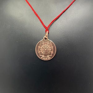 Puede incluir: Un colgante de cobre con un collar de cuerda roja. El colgante presenta un diseño circular con una inscripción en sánscrito y un símbolo en el centro.