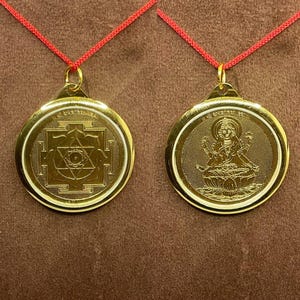 May include: Two gold-colored pendants with red cords. One pendant features a geometric design, the other depicts a seated figure. Both are round with a gold border and a loop for the cord.