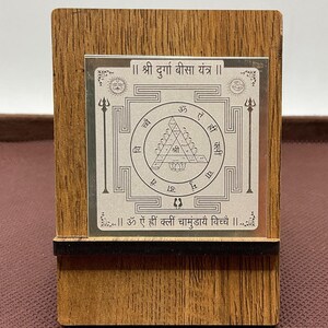 Może przedstawiać: Drewniana ramka z srebrną tabliczką w środku. Tabliczka przedstawia srebrny yantra z tekstem "श्री दुर्गा बीसा यंत्र" i "ॐ ऐं ह्रीं क्लीं चामुंडायै विच्चे".