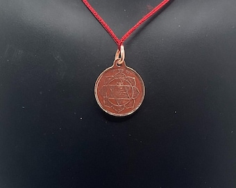 May include: A copper-colored pendant with an intricate geometric design, suspended from a thin red cord. The pendant is circular, featuring a central star within a floral-like border. The necklace is displayed against a dark background.