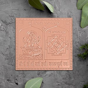 May include: A copper-colored metal yantra with a raised design of a deity sitting on a lotus flower and a yantra with a star inside. The text on the yantra reads "Shree Kamala Pujan Yantra" and "Shree Kamala Yantra" and "Om Aim Hrim Shrim Kleem Hsouh Jagatrasooyai Namah".