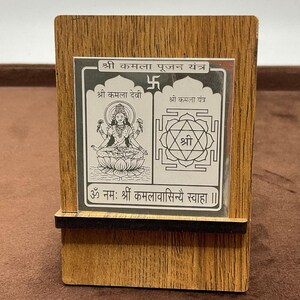 May include: A wooden frame with a silver plaque featuring a deity image and a symbol. The plaque has the text "श्री कमला पूजन यंत्र" at the top, "श्री कमला देवी" on the left, "श्री कमला यंत्र" on the right, and "श्री" in the center. The bottom of the plaque has the text "ॐ नमः श्रीं कमलावासिन्यै स्वाहा ।।"