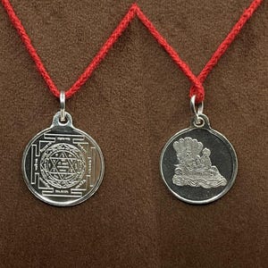 May include: Two silver pendants on red cords. One pendant features an intricate geometric design, while the other depicts a detailed scene with figures. The pendants are round and hang against a brown background.