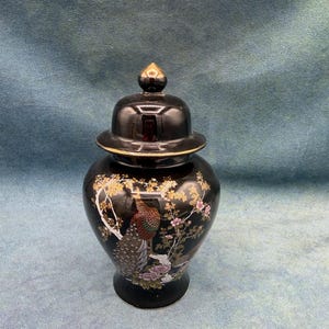 May include: A black ceramic ginger jar with a matching lid featuring a gold-toned finial. The body of the jar is decorated with a detailed illustration of a peacock perched on a branch surrounded by pink and white floral blossoms and foliage.