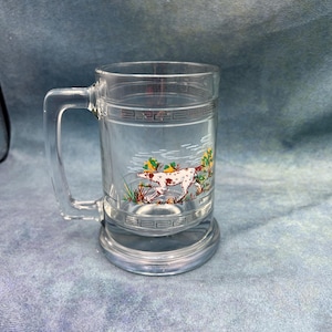 May include: A clear glass beer mug with a handle. The mug has a design of a dog running through a field with trees and bushes. The design is in shades of brown, white, green, and yellow.