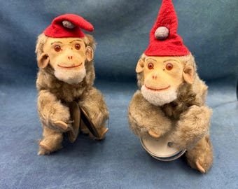 May include: Two vintage toy monkeys with brown fur, red hats, and smiling faces. One monkey holds cymbals, the other a drum. The toys are set against a blue background.