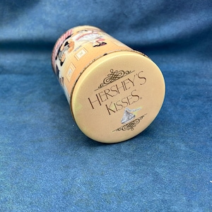 May include: A round, cream-colored tin with a brown design featuring a woman in a red dress and the text "HERSHEY'S KISSES."