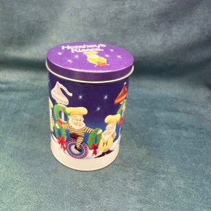 May include: A purple and white tin with a Christmas theme. The tin has a picture of three elves on it. The text on the tin reads "Hershey's Kisses".
