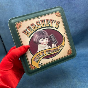 May include: A green and gold tin with the words "Hershey's Milk Chocolate" and a picture of a young girl holding a Hershey's chocolate bar.