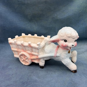 May include: A ceramic planter shaped like a white lamb pulling a small, rectangular, fence-style cart. The lamb has pink-tinted cheeks, ears, and hooves, with a small gold-colored bell around its neck. The cart is white with pink accents.