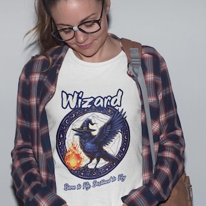 May include: White t-shirt with a blue raven wearing a witch's hat and holding a flame. The raven is surrounded by a circle with stars and a pentagram. The text "Wizard" is above the raven and "Born to Fly, Destined to Fly" is below the raven.