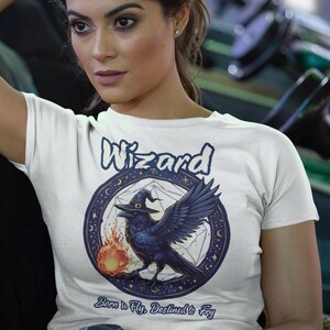 May include: White t-shirt with a graphic of a black raven wearing a wizard hat and holding a flame. The graphic is surrounded by a circle with the words "Wizard" and "Born to Fly, Destined to Fly".