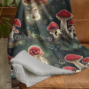 May include: A cozy throw blanket featuring a whimsical design of red and white spotted mushrooms against a dark green background. The blanket is draped over a brown leather couch, showcasing its soft, plush texture and detailed illustration.