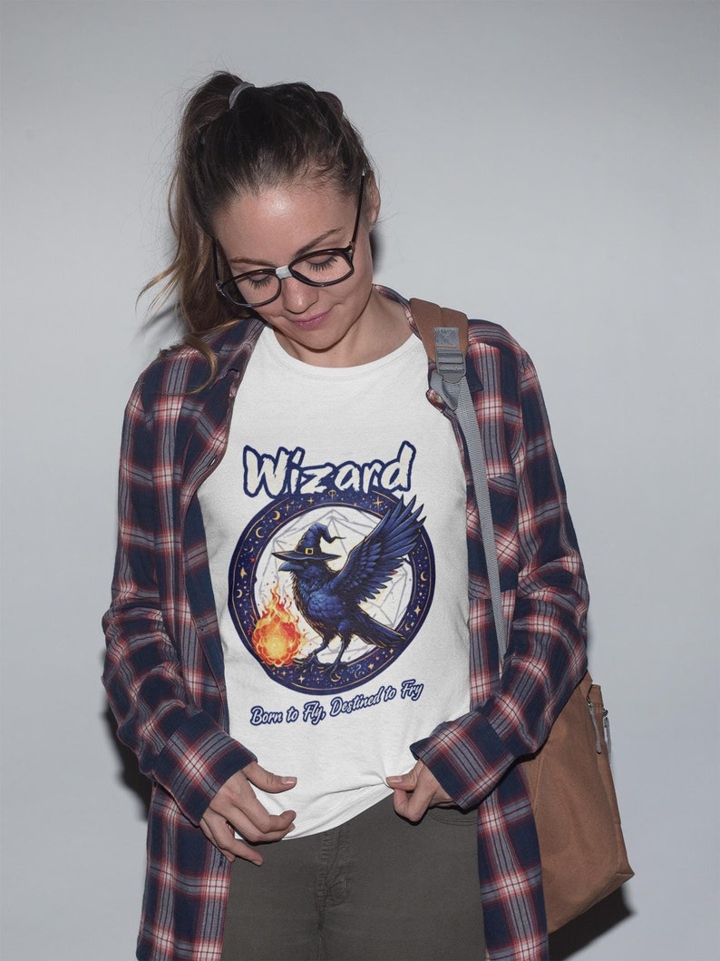 May include: White t-shirt with a blue raven wearing a witch's hat and holding a flame. The raven is surrounded by a circle with stars and a pentagram. The text "Wizard" is above the raven and "Born to Fly, Destined to Fly" is below the raven.