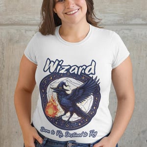 May include: White t-shirt with a black raven holding a flame in a blue and white circle with the word "Wizard" above it and the text "Born to Fly, Destined to Fly" below it.