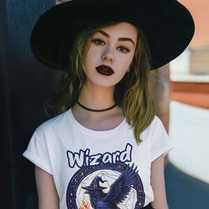 May include: A woman wearing a black witch hat and a white t-shirt with a graphic of a raven wearing a witch hat and the word "Wizard" printed on it.