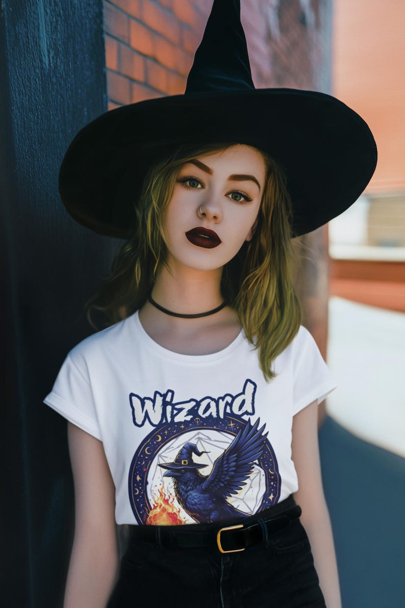 May include: A woman wearing a black witch hat and a white t-shirt with a graphic of a raven wearing a witch hat and the word "Wizard" printed on it.