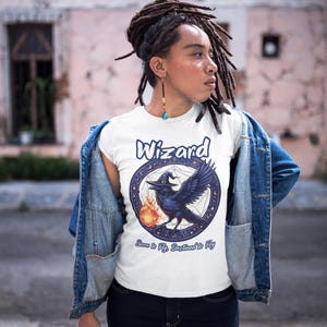 May include: White t-shirt with a blue and white graphic of a raven wearing a witch's hat and holding a fireball. The graphic is surrounded by a circle of stars and the text "Wizard Born to Fly, Destined to Fly".