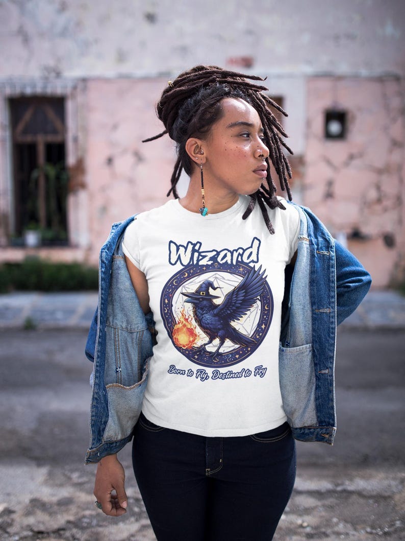 May include: White t-shirt with a blue and white graphic of a raven wearing a witch's hat and holding a fireball. The graphic is surrounded by a circle of stars and the text "Wizard Born to Fly, Destined to Fly".