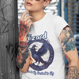May include: White t-shirt with a blue and white graphic of a raven wearing a witch's hat. The graphic is surrounded by a circle of stars and a pentagram. The text "Wizard" is above the graphic and "Born to Fly. Destined to Fry" is below the graphic.