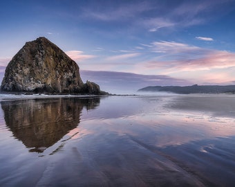 May include: A large rock formation, known as Haystack Rock, stands tall on a sandy beach. The rock is reflected in the calm, still water at low tide. The sky is a soft blue with wispy clouds, and the horizon is a pale pink.