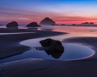 May include: A scenic view of a rocky beach at sunset. The sky is a vibrant mix of pink, purple, and blue, reflecting in the calm water. A small pool of water has formed on the sand, creating a natural mirror reflecting the sky.