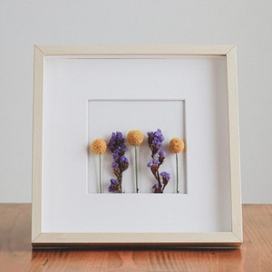 Peut inclure: Une œuvre d'art florale encadrée avec des fleurs séchées. L'arrangement comprend de petites fleurs rondes orange et des brins violets sur fond blanc. Le cadre est en bois clair, créant une esthétique minimaliste.