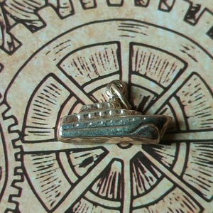 Peut inclure: Un charm en argent en forme de bateau avec des balustrades détaillées et une petite boucle d'attache. Le charm est posé sur un fond beige avec un motif d'engrenage.