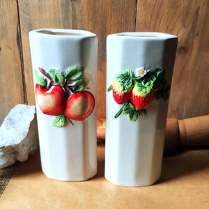 May include: Two white ceramic vases with a faceted design. One vase features a pair of red apples with green leaves, and the other has red strawberries with green leaves and a small white flower. The vases are decorative.
