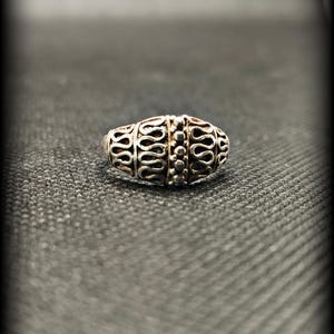 Könnte beinhalten: Ein silberfarbener Ring mit ovaler Form und aufwendigem filigranem Design. Der Ring weist eine Reihe von verbundenen, wellenförmigen Mustern auf, mit einer Reihe kleiner, erhabener Punkte in der Mitte. Der Ring ist vor einem dunklen Hintergrund platziert.