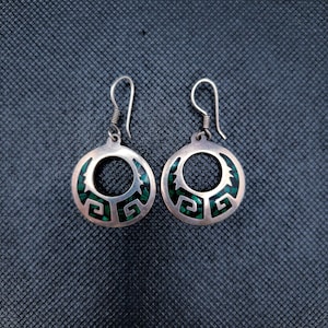 Peut inclure: Une paire de boucles d'oreilles argentées avec un motif circulaire. Les boucles d'oreilles présentent un cercle central ouvert avec un motif géométrique et des incrustations de pierres vertes. Fermeture à crochet.