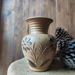 Könnte beinhalten: Eine beige Keramikvase mit einem floralen Design, das in die Oberfläche geschnitzt ist. Die Vase hat einen breiten Boden, einen abgerundeten Körper und einen ausgestellten Rand. Das florale Design zeigt stilisierte Blumen und Blätter. Die Vase steht auf einer Jutefläche.