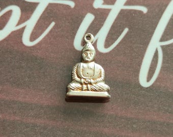 Może przedstawiać: Mały, złoty wisiorek z Buddą. Budda przedstawiony jest w pozycji siedzącej medytacji, ze skomplikowanymi detalami w ubraniach i rysach twarzy. Wisiorek ma małą pętlę na górze do przymocowania do łańcuszka lub sznurka.